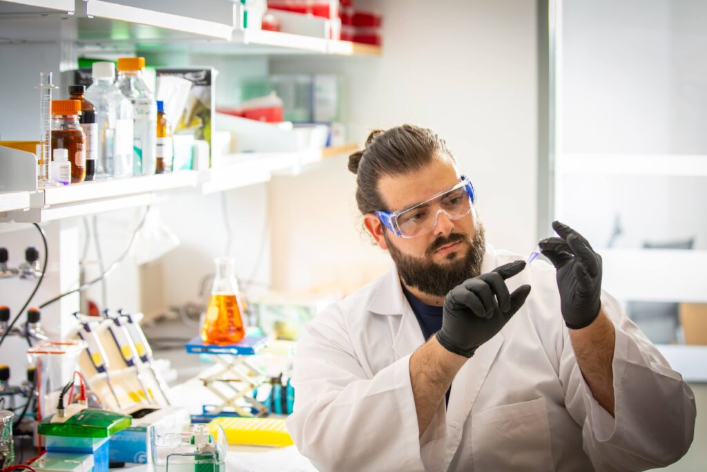 A lab member in a lab coat and safety goggles holding a sample at the bench